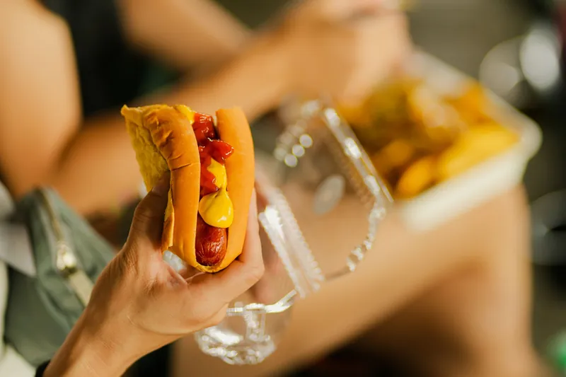 A hot dog with ketchup and mustard held in one hand, with fries behind, at an outdoor food stall